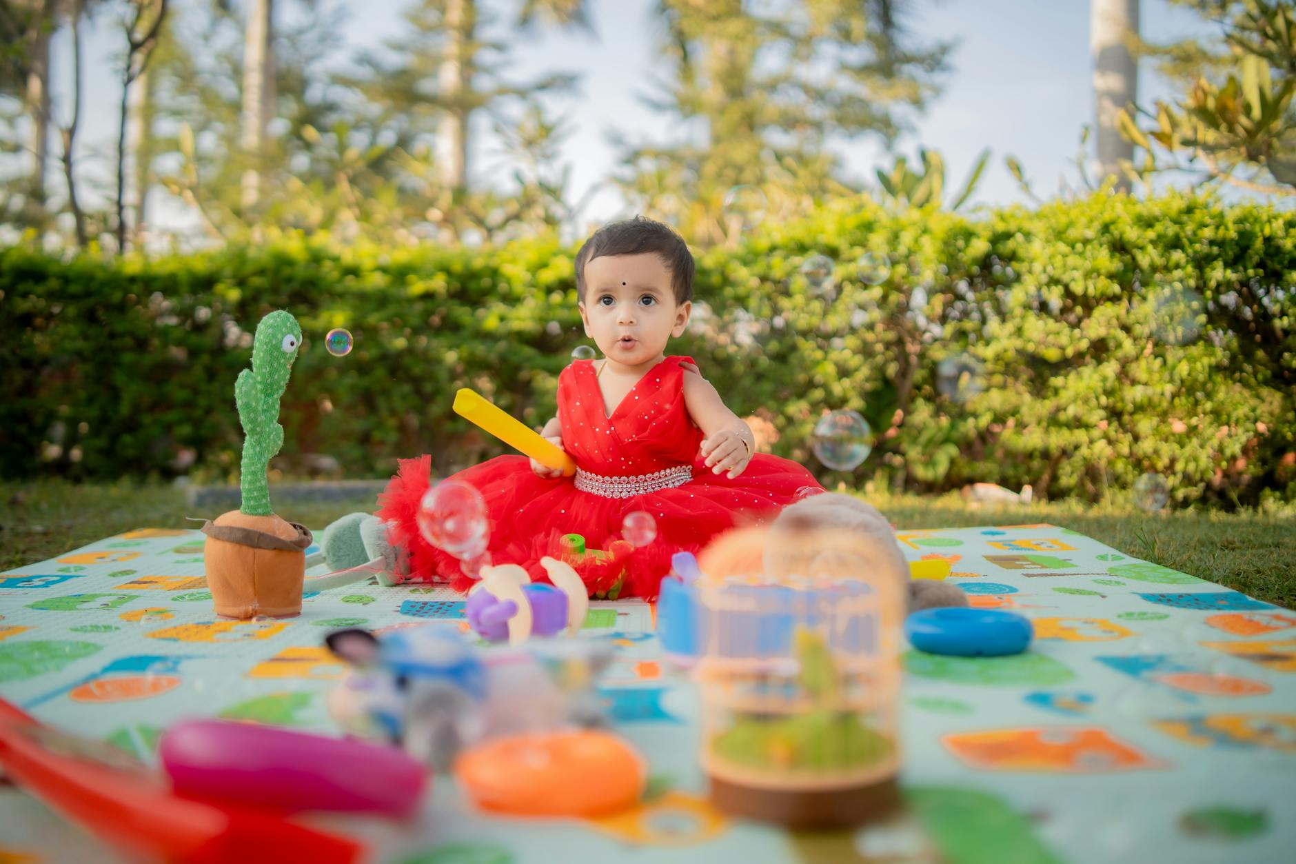 Adorable baby in a red dress playing with toys on a colorful mat outdoors.