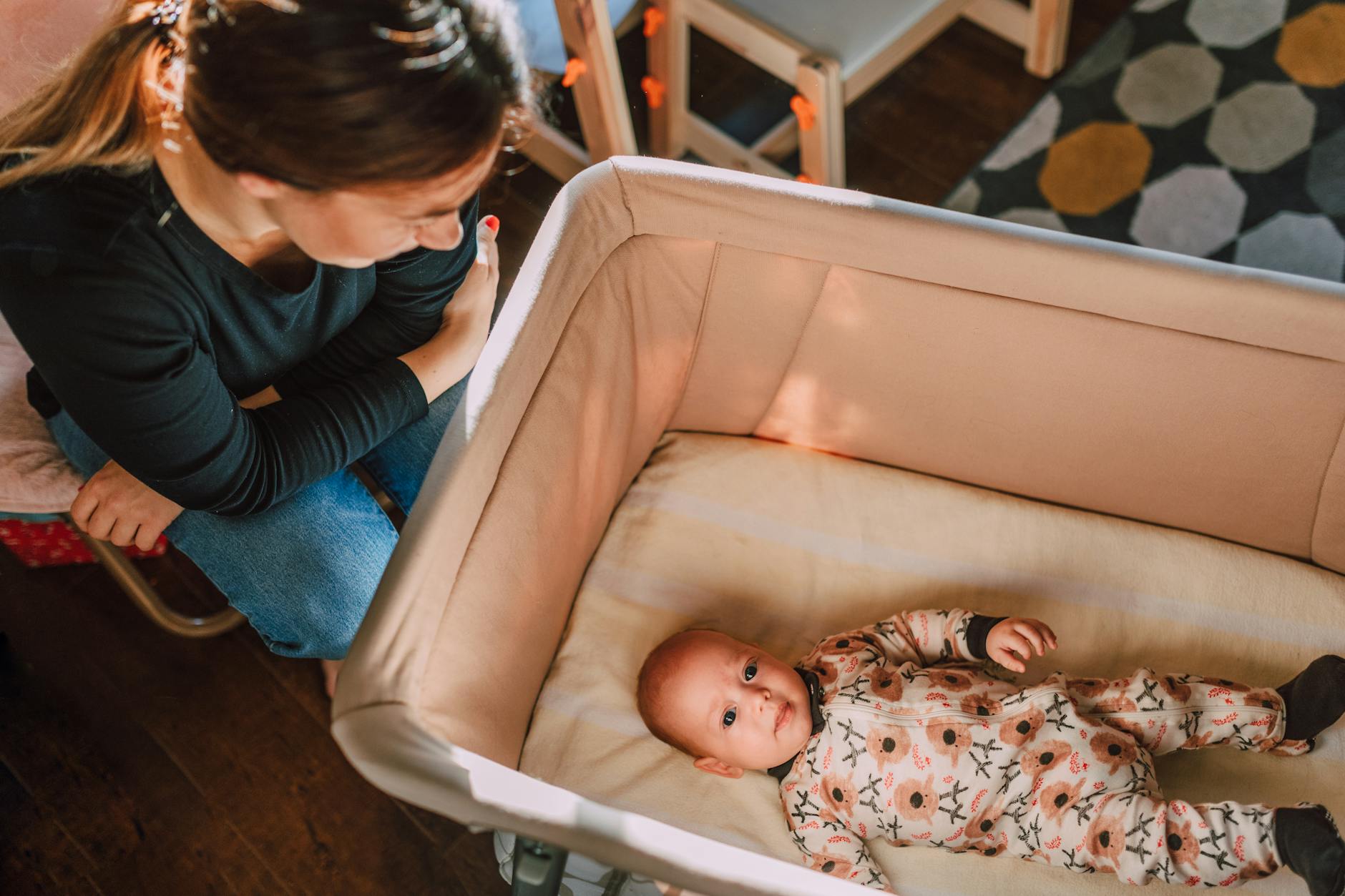 A mother lovingly watches over her baby in a crib from an overhead view.