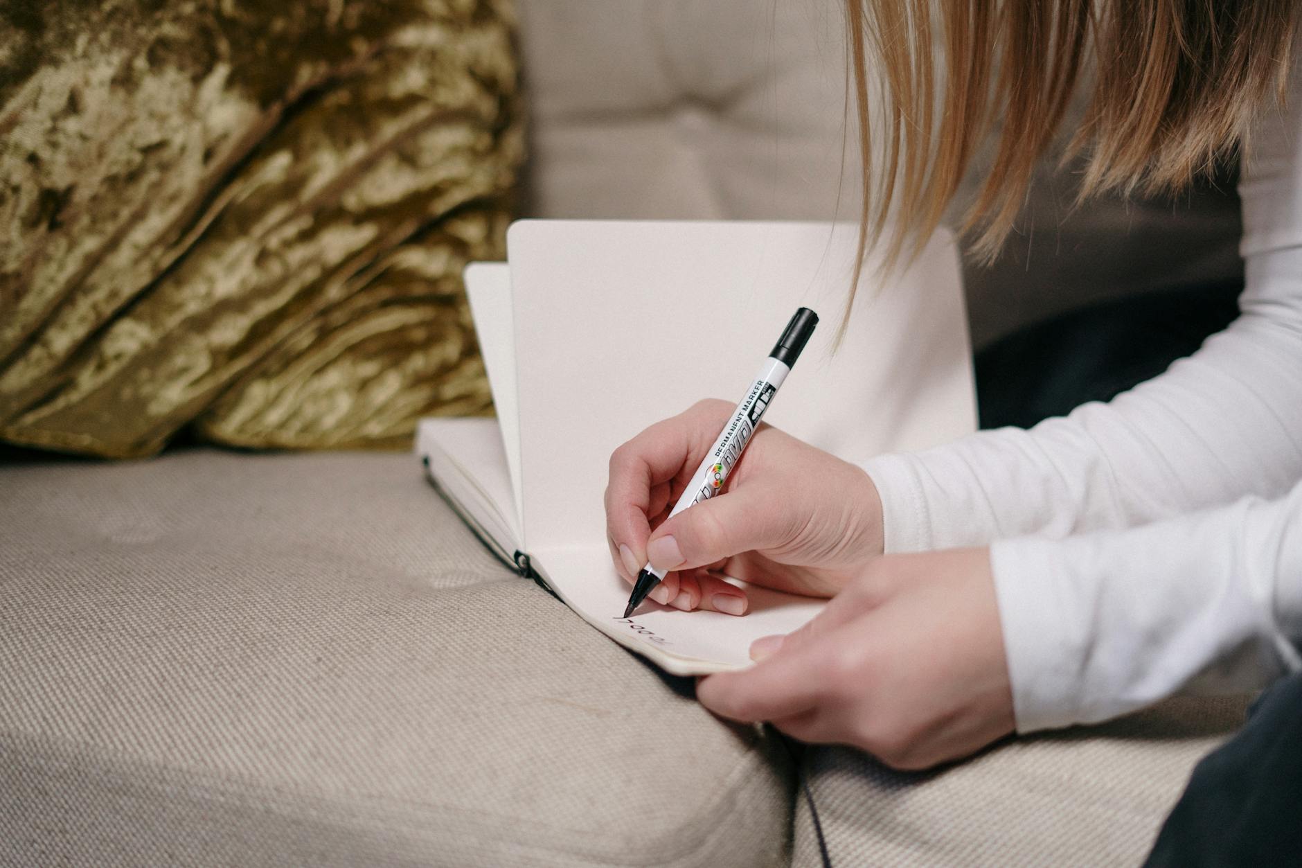 Close-up of a person writing in a notebook with a pen, sitting on a couch indoors.