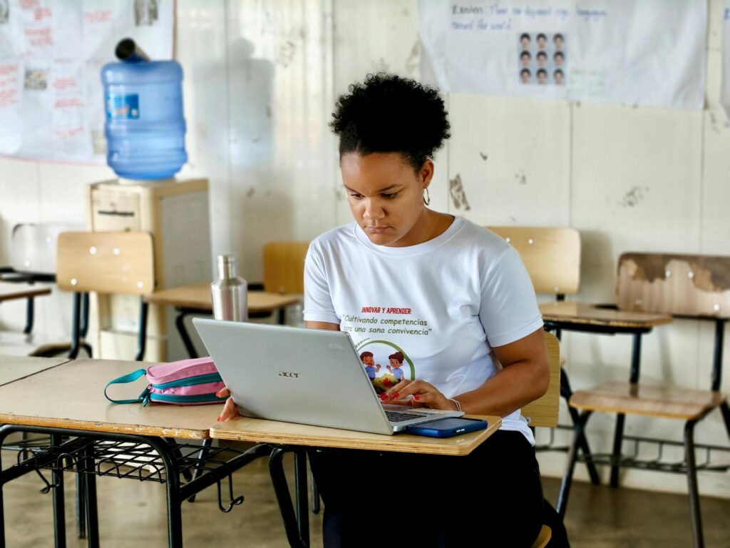 Woman focused on a laptop in a classroom environment, surrounded by desks and educational posters.