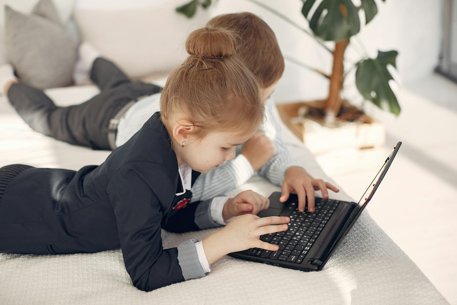 Focused boy and girl in formal outfits typing on netbook while resting on cozy bed during free time at home