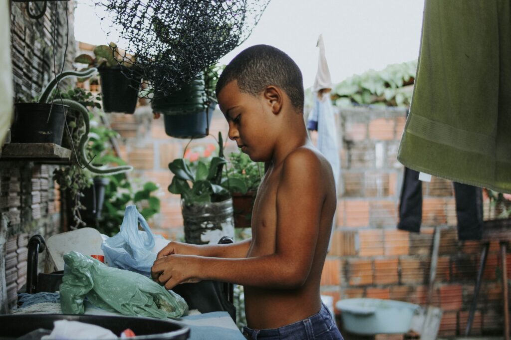 A boy stands in a rural yard, organizing items on a table surrounded by plants.