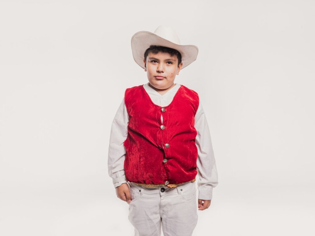 Portrait of a young boy wearing traditional Mexican attire, featuring a red vest and white hat, against a studio white background.