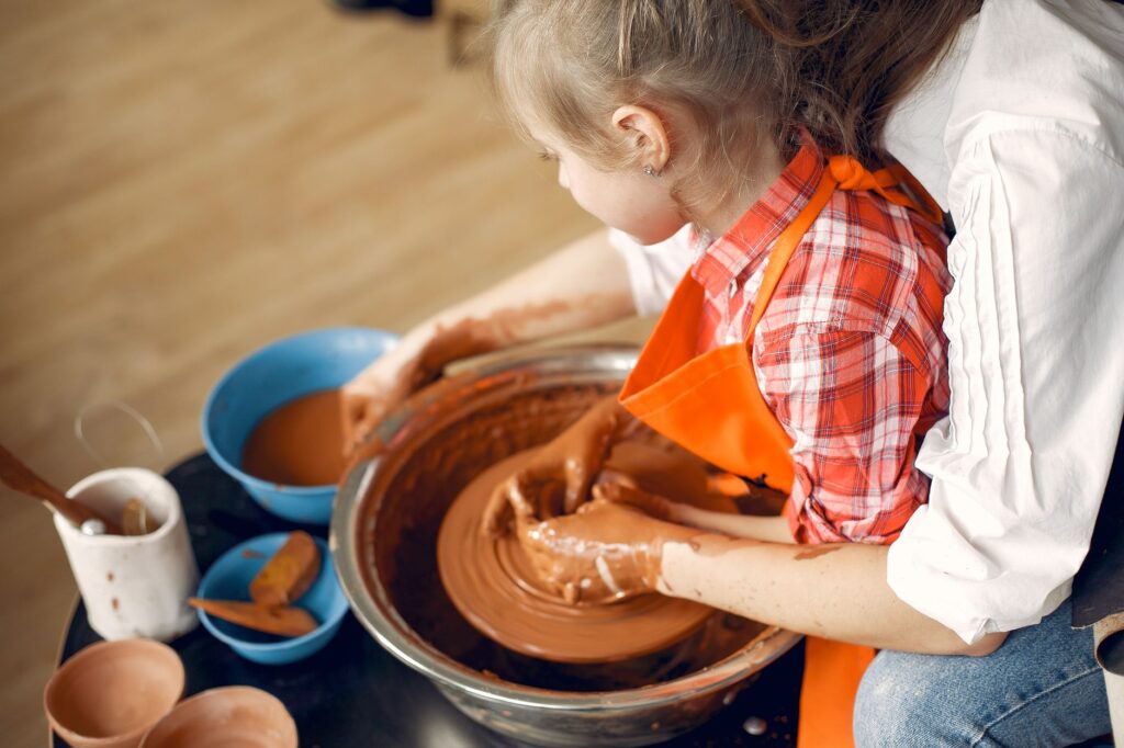 From above side view crop anonymous female potter with little daughter in apron making clay product together using pottery wheel