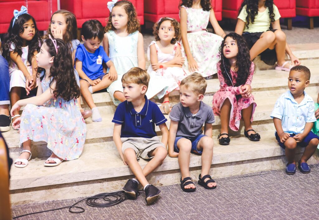 Children attentively sit on marble steps indoors, dressed in colorful summer clothing.
