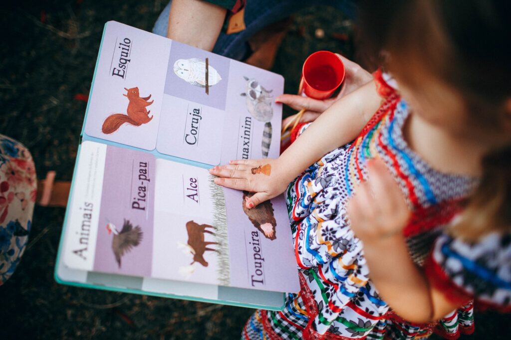 From above of little girl with temporary tattoo on hand in vivid summer dress reading colorful book with big pictures near crop woman in park on daytime