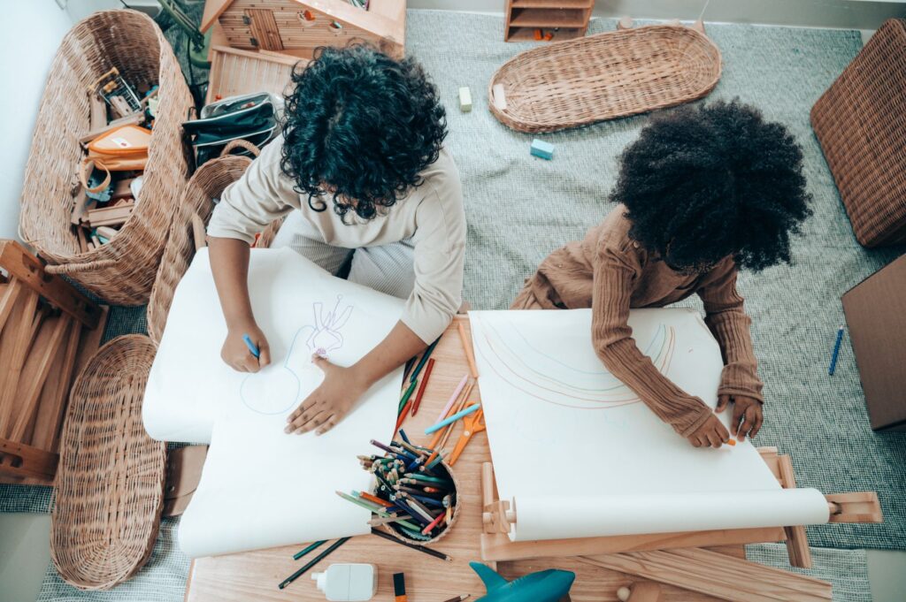 Top view of unrecognizable black children with Afro hairstyle sitting at wooden table with paper roll while drawing on paper sheets with different pencils in flat