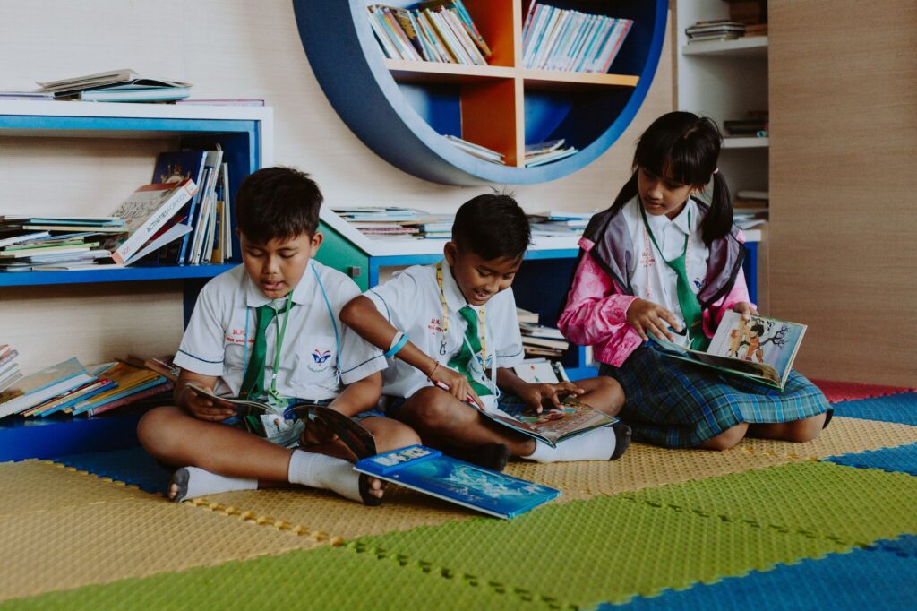 Three children in school uniforms reading books together in a vibrant library setting.