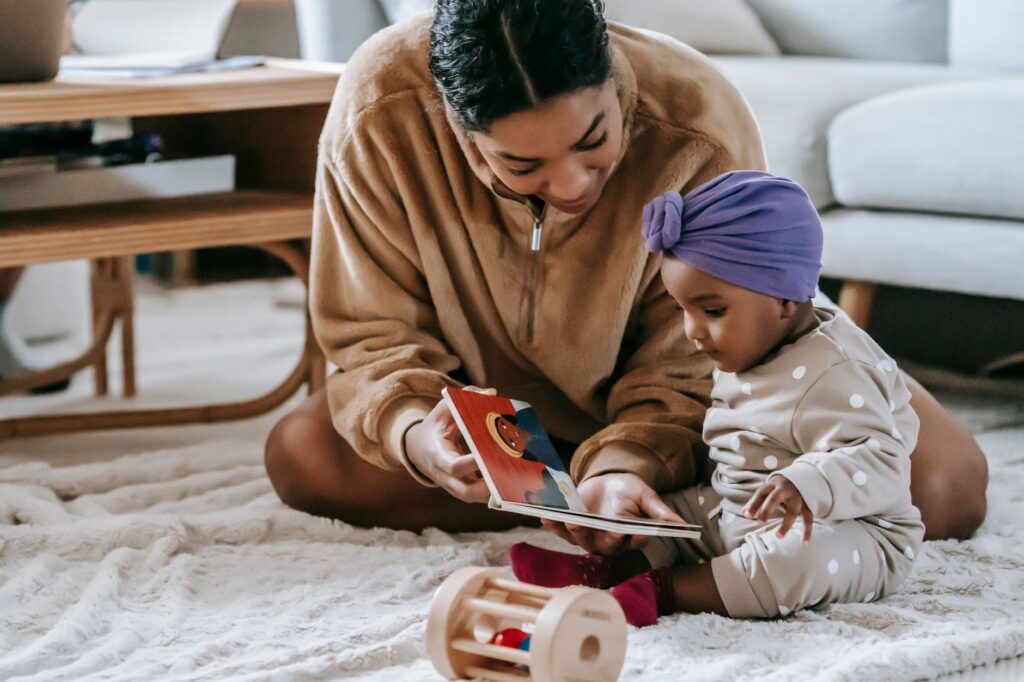 Crop African American mother demonstrating book to little black daughter in headwear while sitting on rug with toy at home