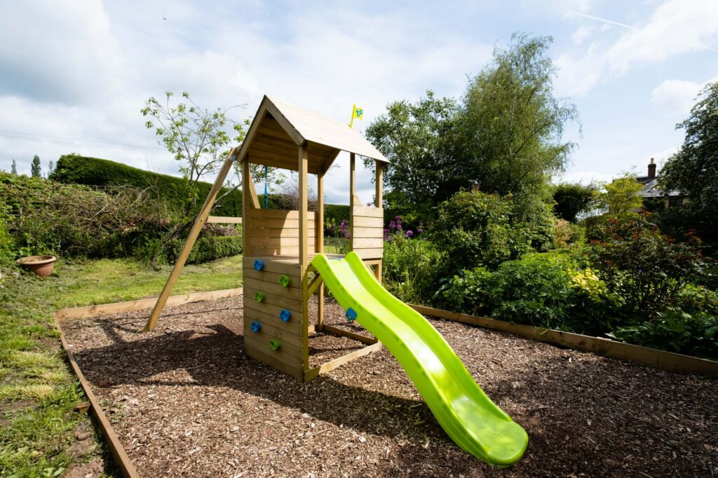 Wooden play structure with slide in a lush garden on a sunny day.