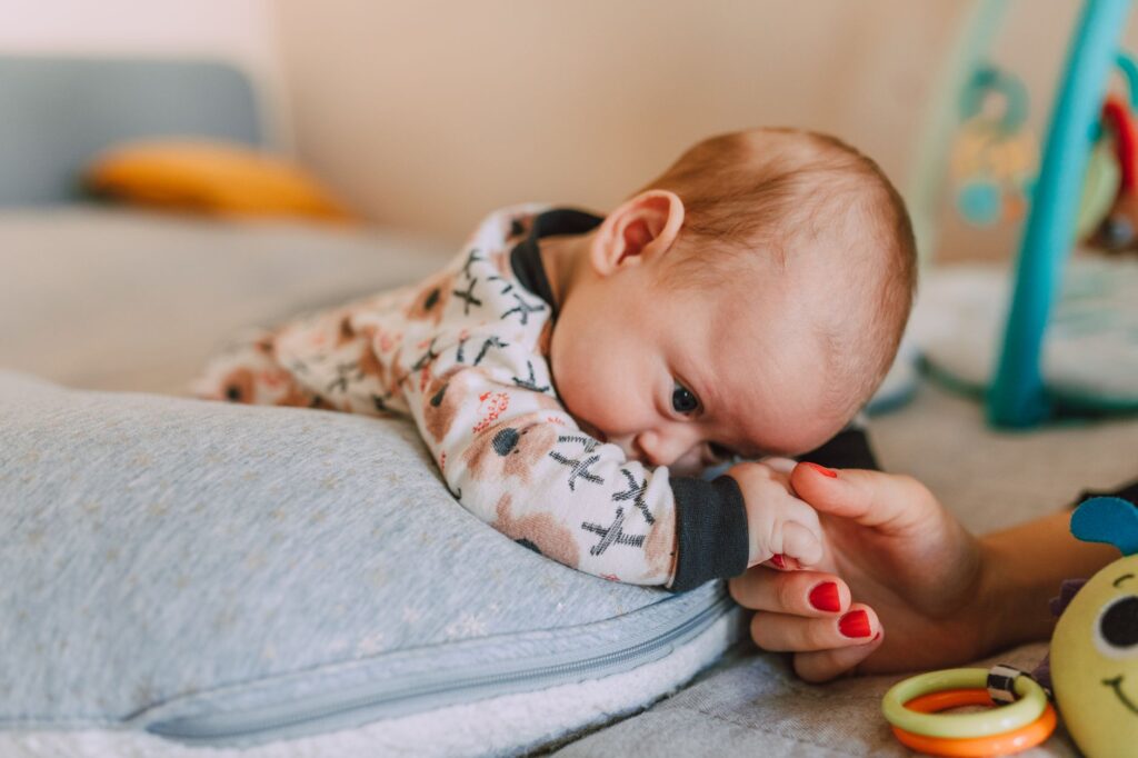 A baby lies on a cushion while affectionately holding an adult's hand indoors.