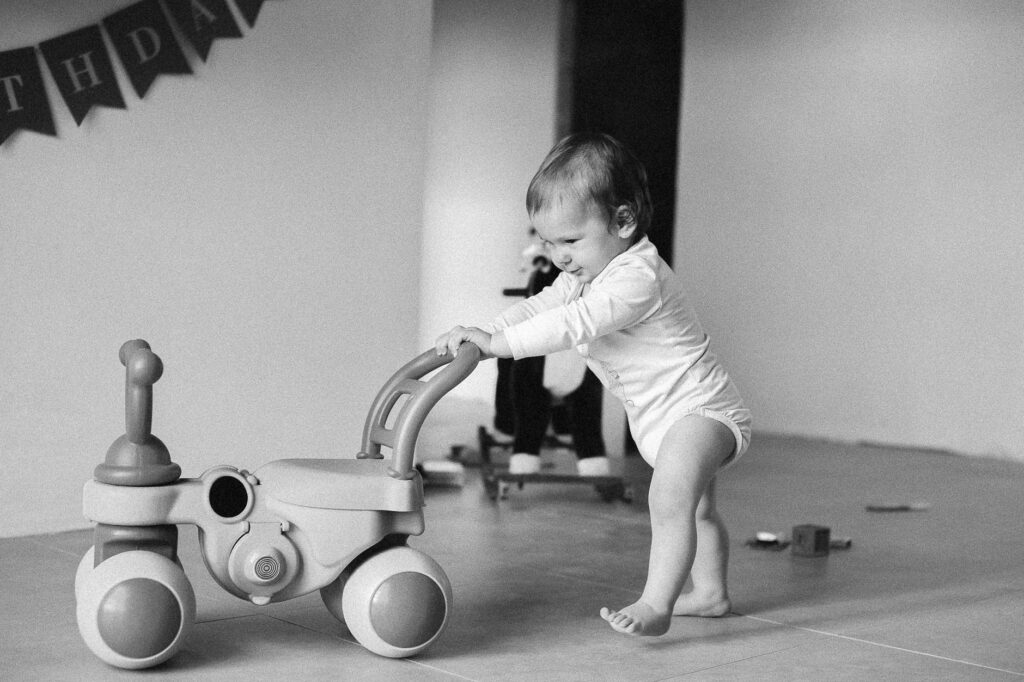A black-and-white photo of a toddler pushing a toy indoors, capturing youth and innocence.
