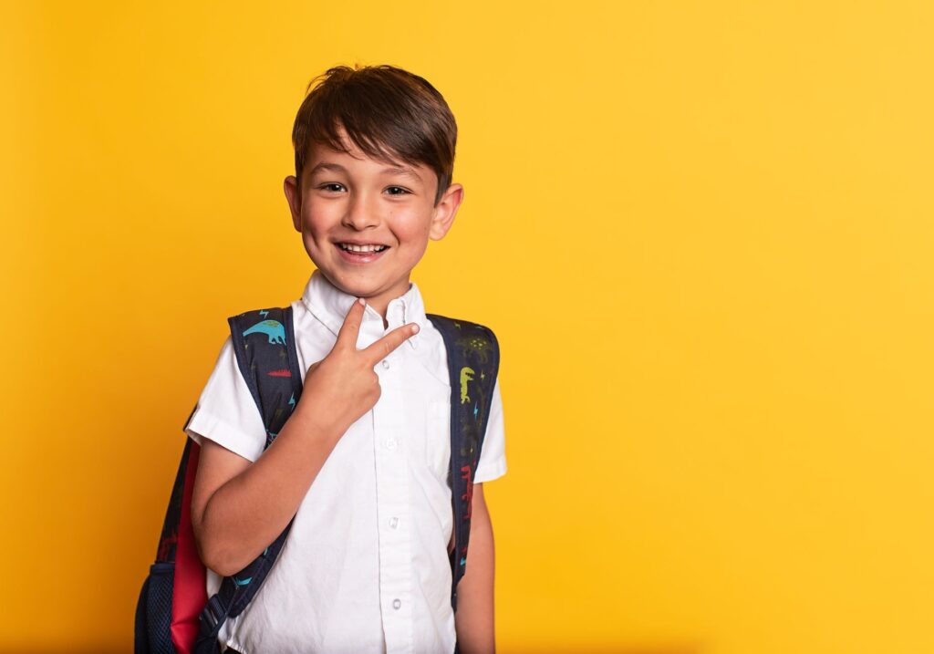 Cheerful boy in white shirt with backpack poses on yellow background, ready for school.