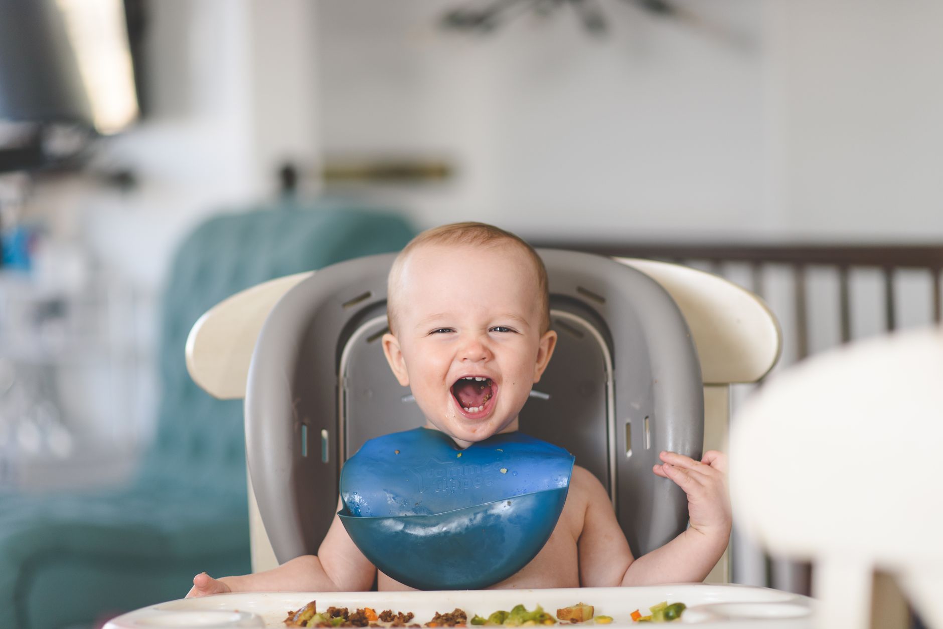 Happy baby laughing in a high chair during mealtime, wearing a blue bib.