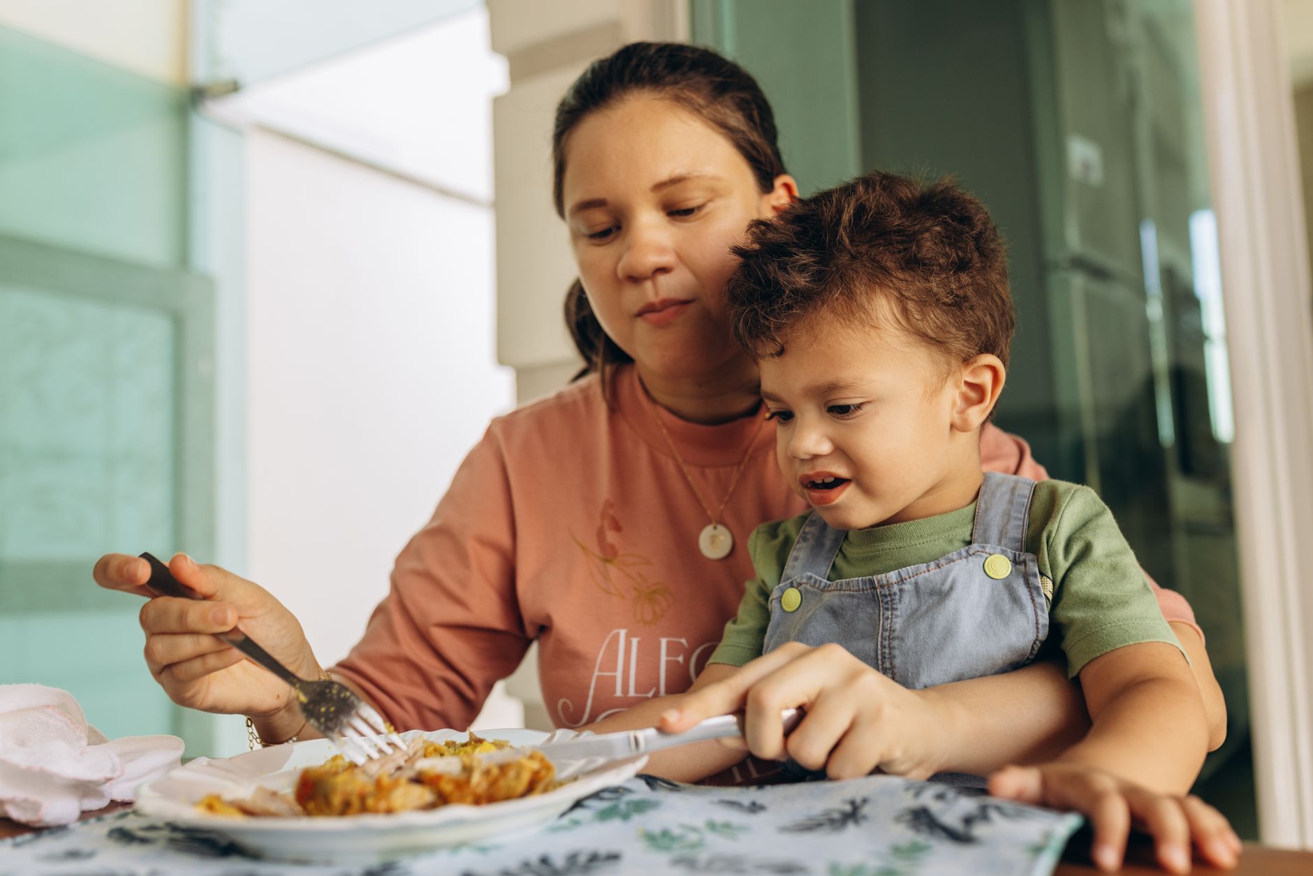 A mother helps her child eat at the kitchen table, fostering family bonding.