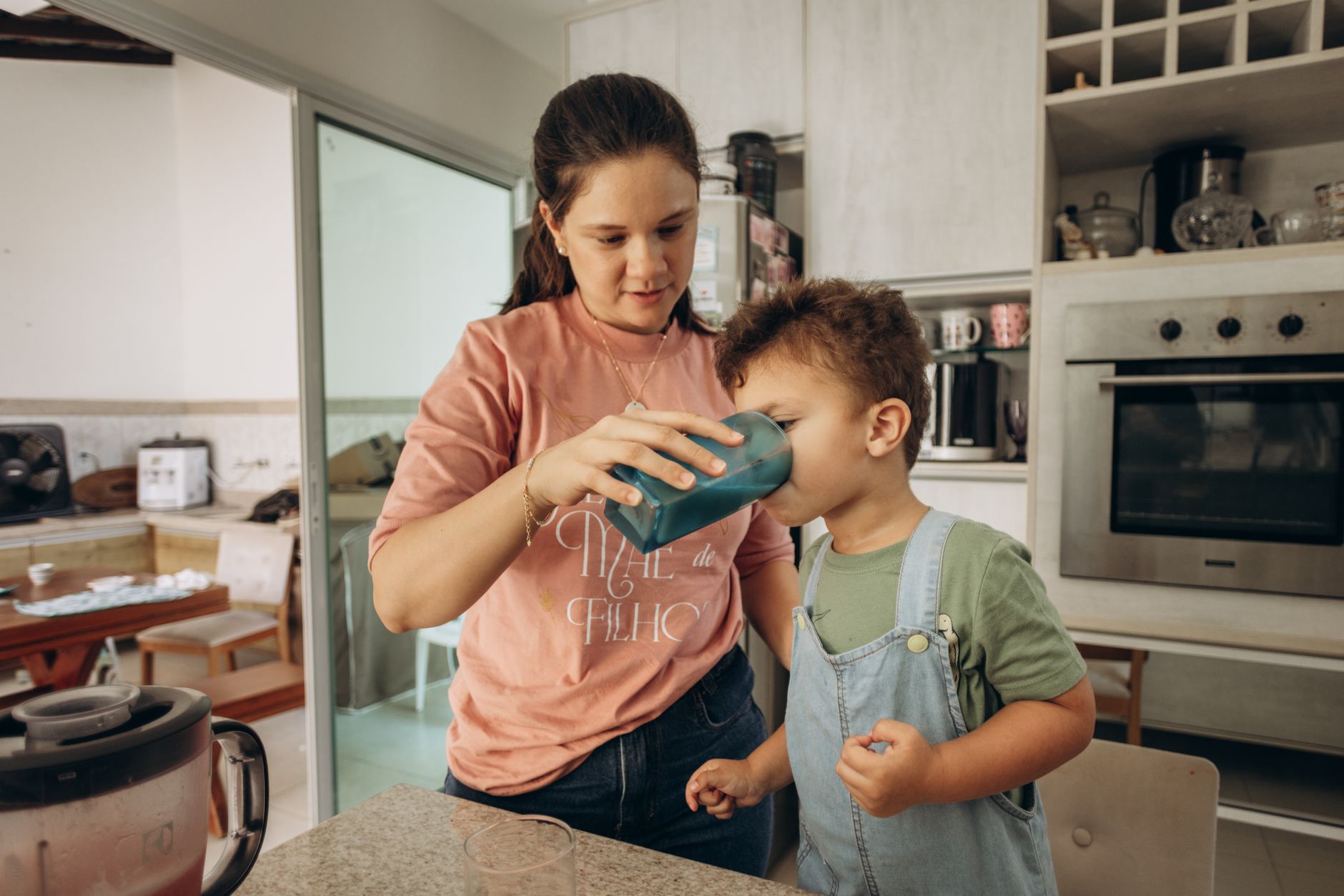 A mother assists her son drinking from a glass in a cozy kitchen setting.