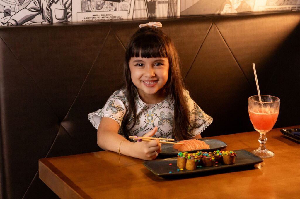 A young girl smiling while eating sushi at a restaurant with a colorful drink.