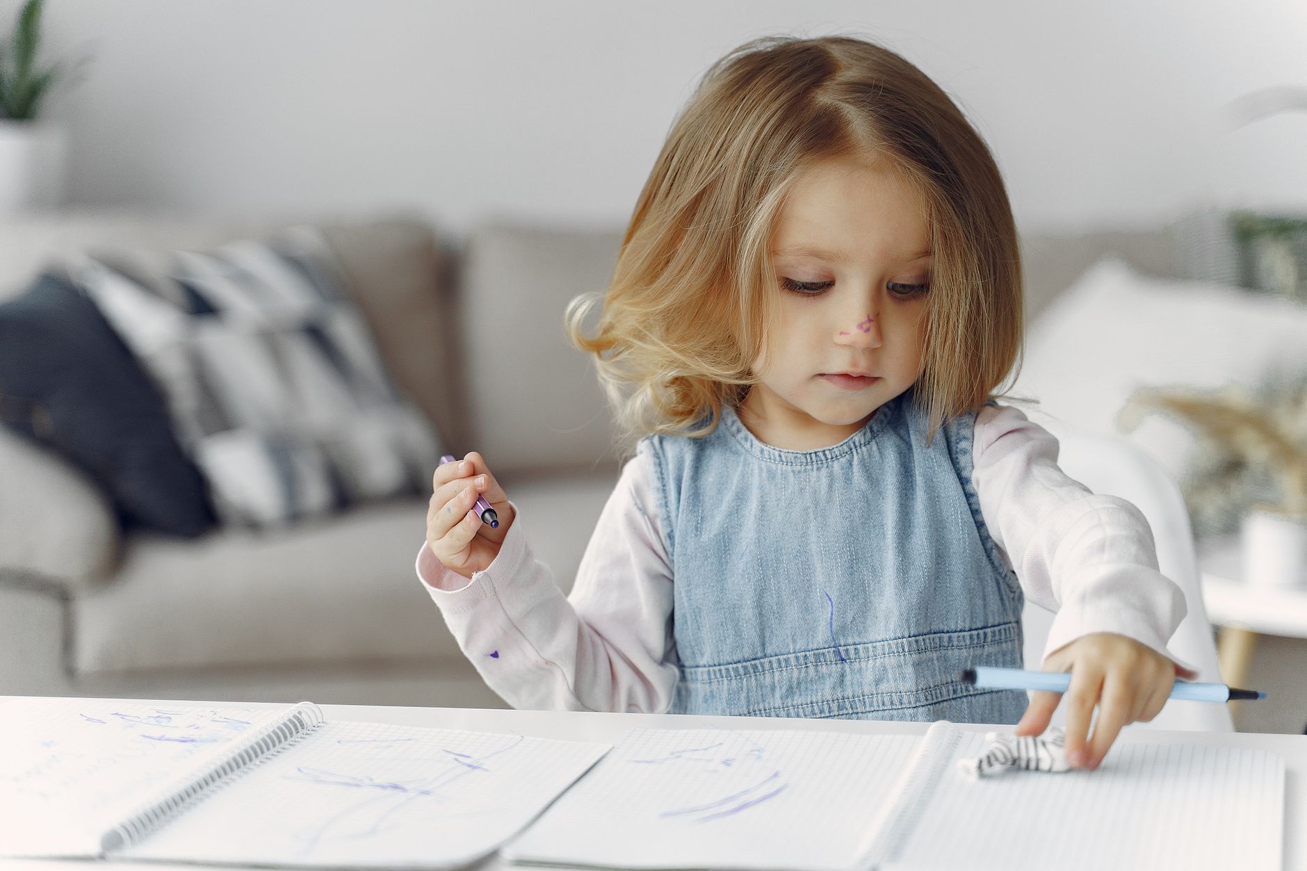 A young girl sits at a table, creatively drawing in her notebook with colored pens, indoors.
