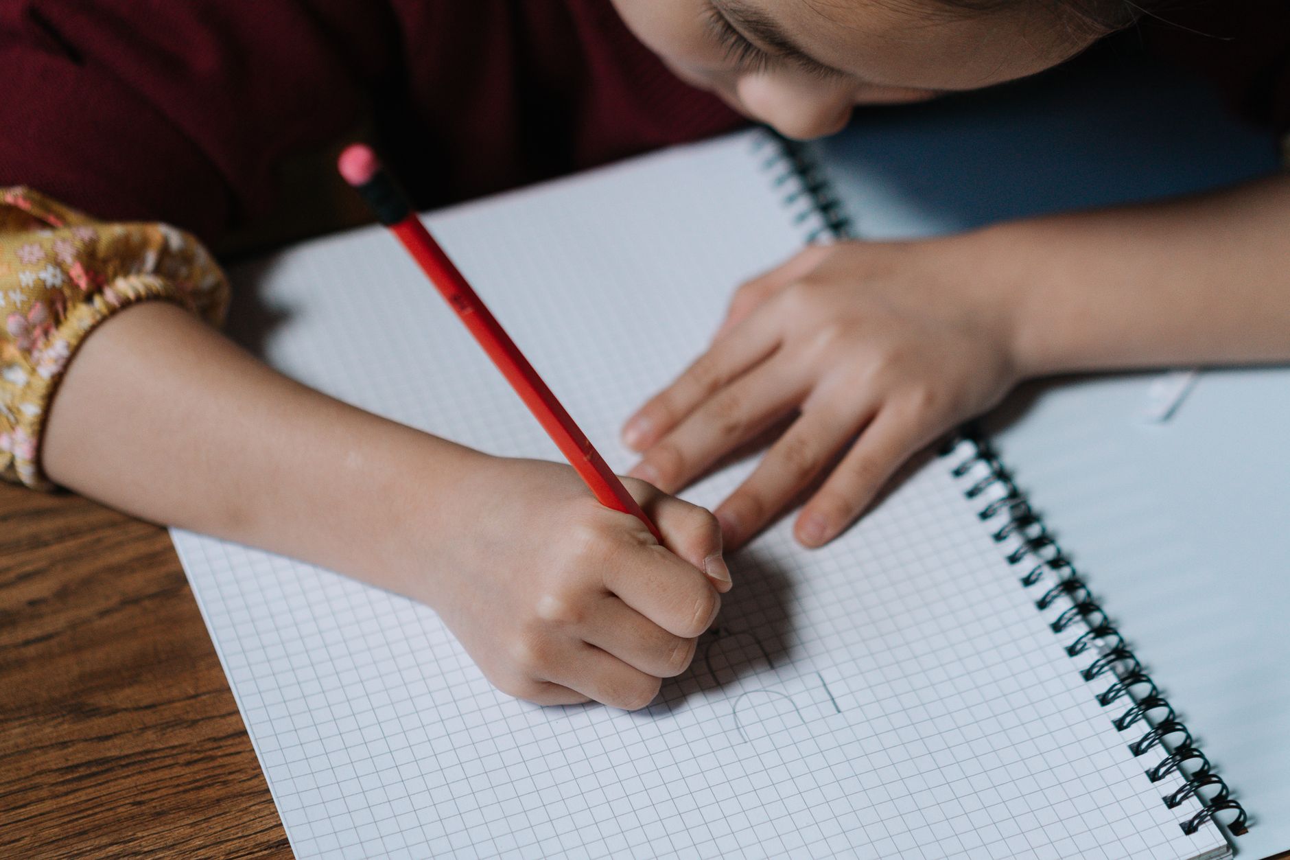 A child writes in a spiral notebook with a red pencil on a wooden desk.