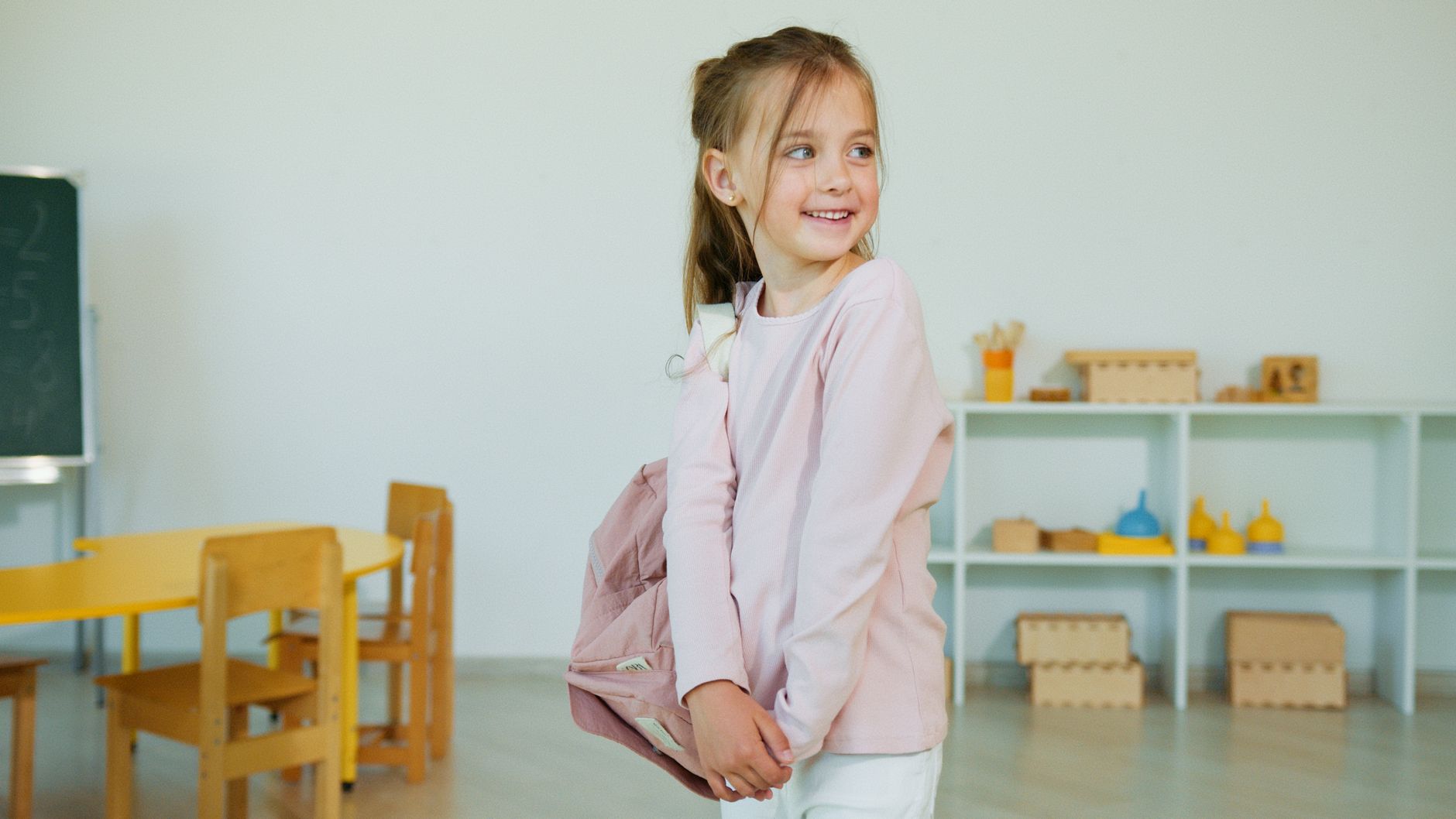 A joyful girl in a classroom setting, wearing a light pink shirt and holding a backpack.