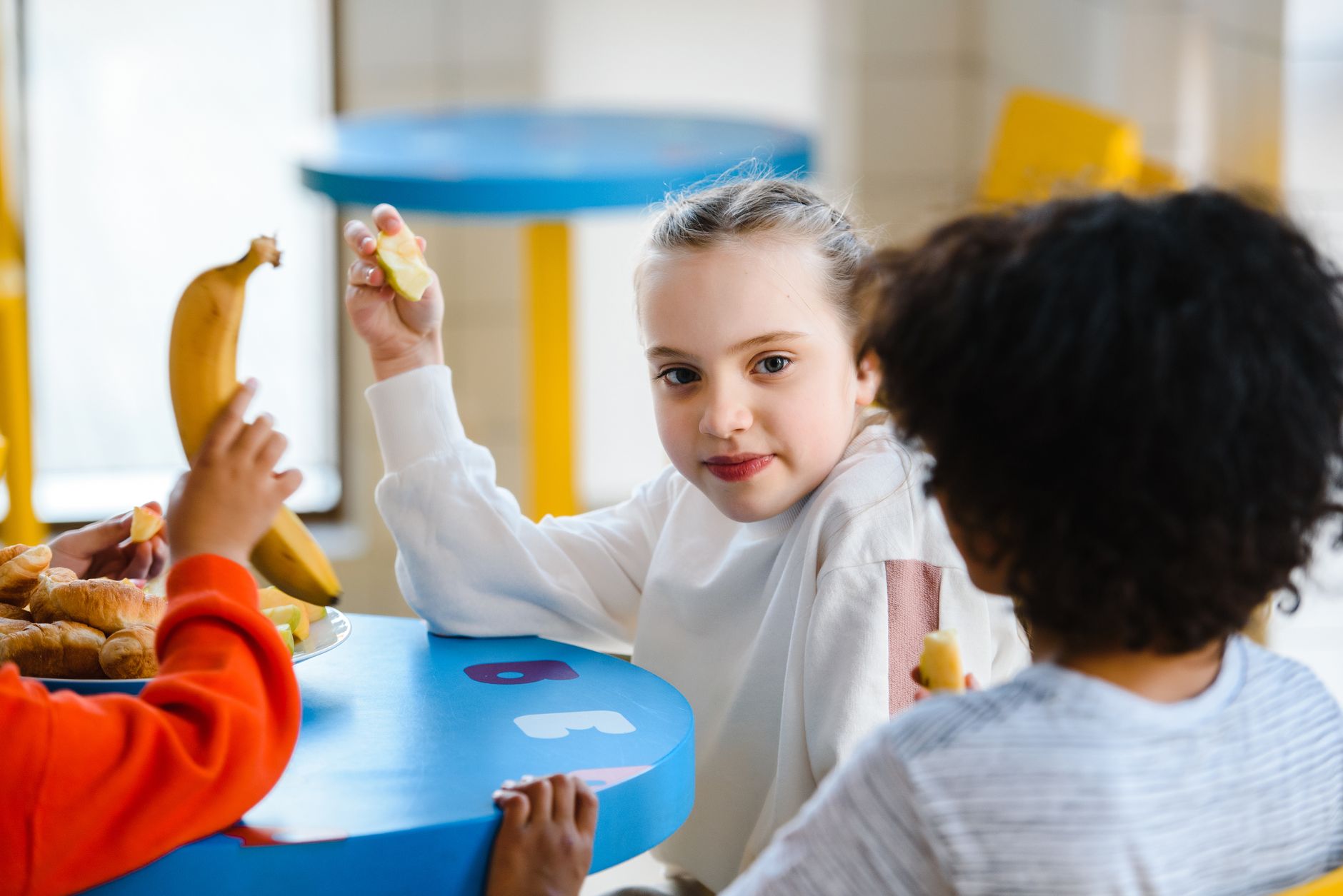 Three children sitting at a table enjoying bananas and pastries during snack time.