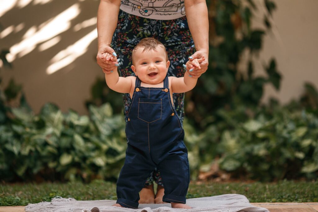 Cheerful baby in overalls being supported while taking first steps outdoors.