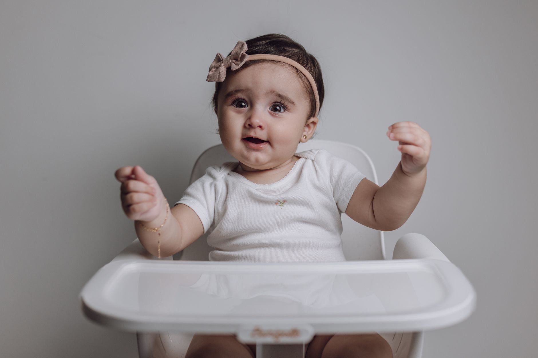 Adorable baby girl with a headband sitting in a white high chair, smiling joyfully.